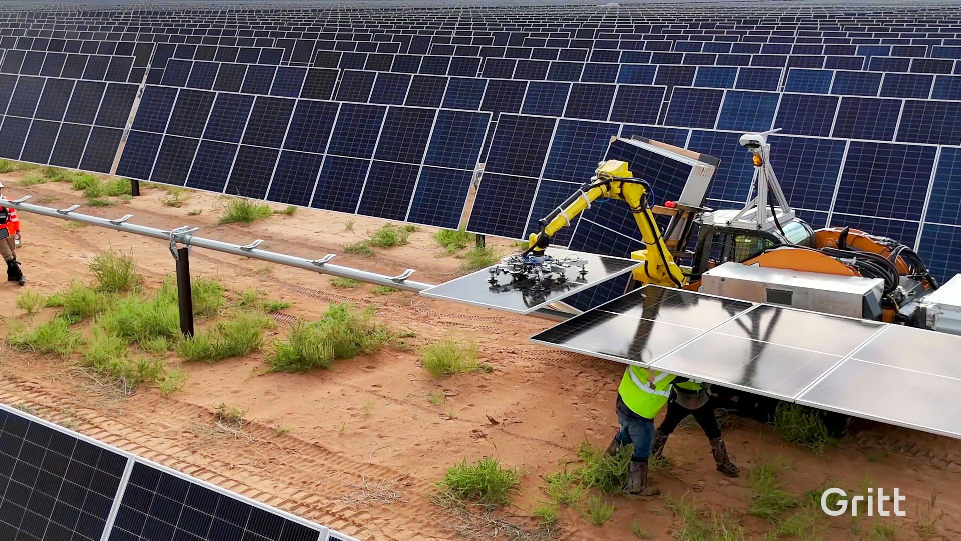 A Gritt robotic arm lowering a solar panel into a row on an active desert solar farm, a worker in safety gear alongside