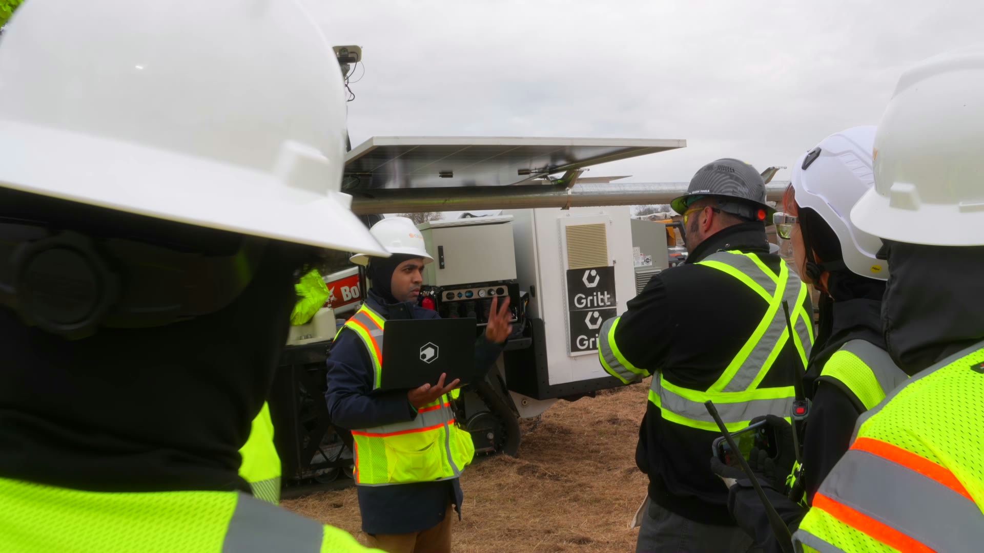 A Gritt crew in hard hats and hi-vis gathered around the machine on a jobsite, one presenting on a laptop